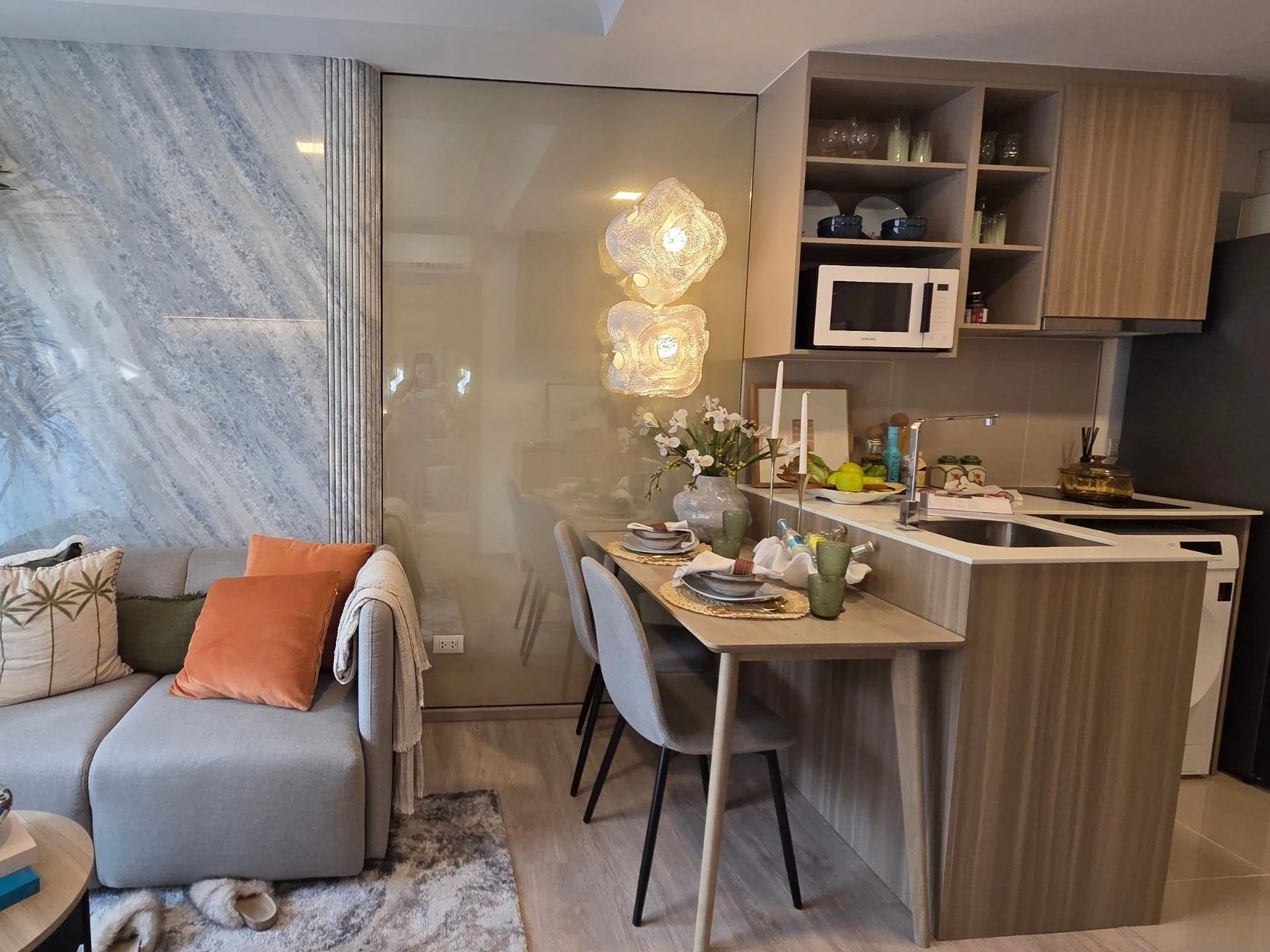 Open-plan kitchen and dining area with grey sofa and orange cushions in a 2-bedroom apartment at Rhea, Surin, Thailand